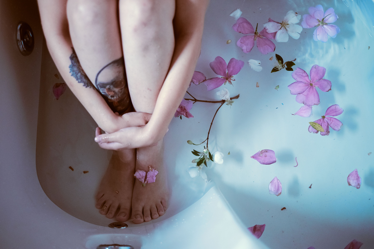 A pair of feet and lower legs submerged in a clear bathtub filled with water, surrounded by floating pink flower petals, symbolizing female pleasure.