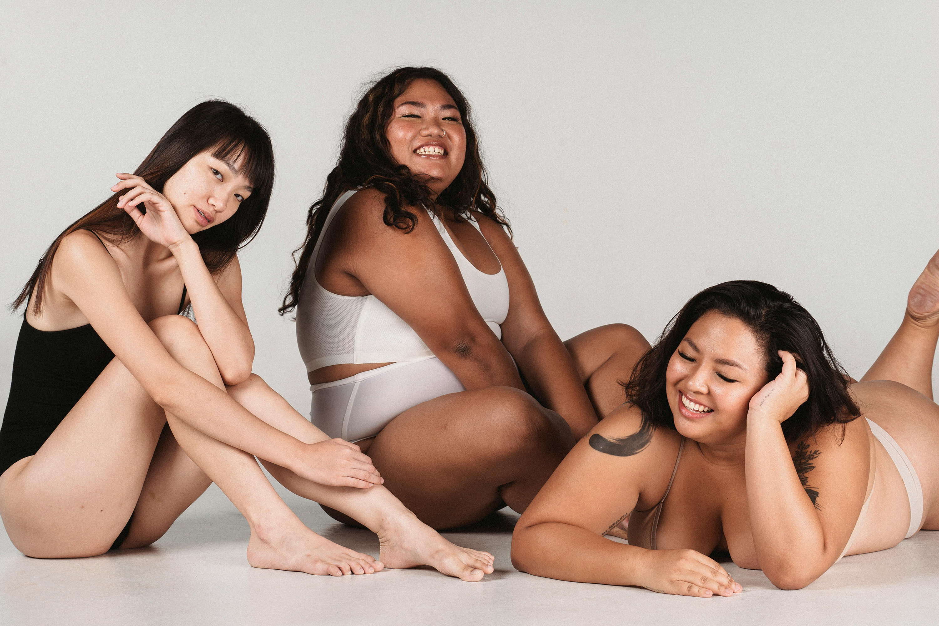 Three smiling women of different body types posing together in simple black and white garments against a plain background.