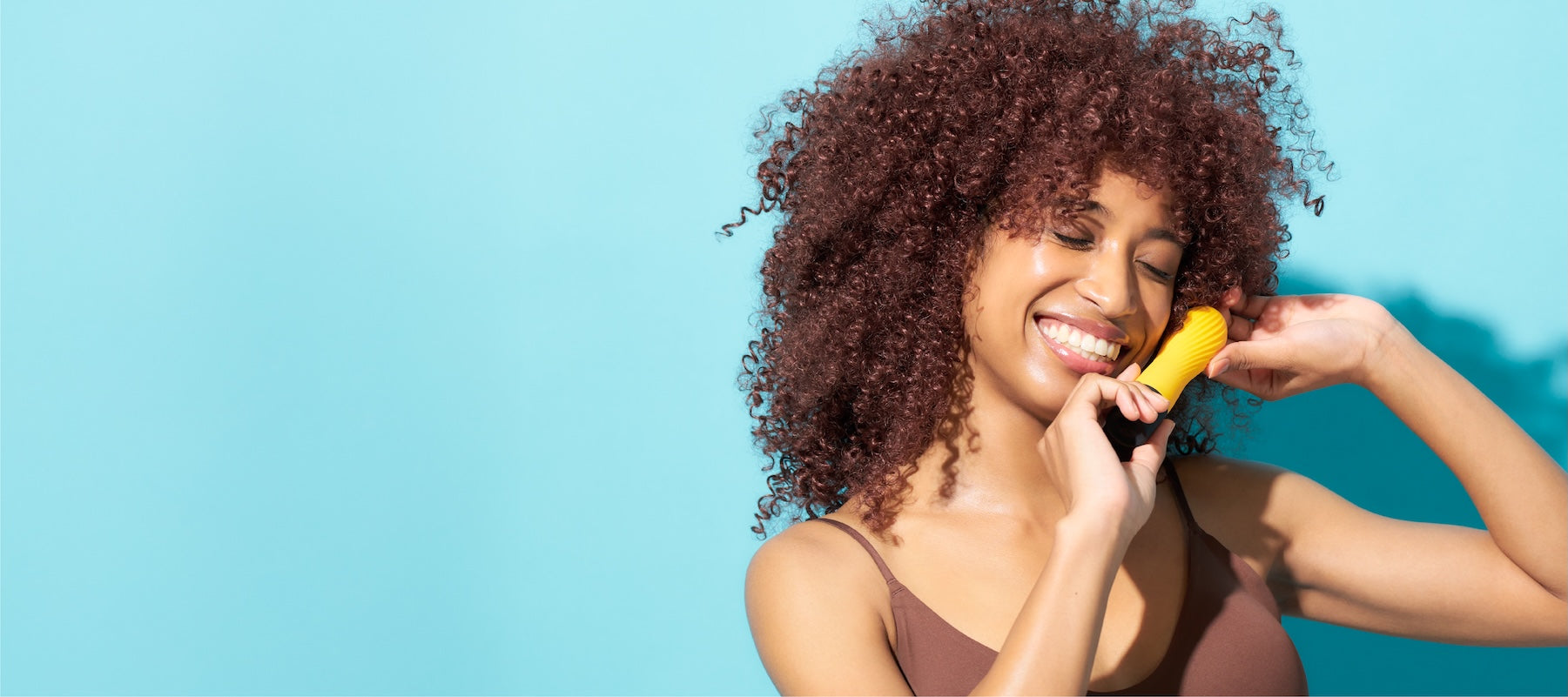 Woman with curly hair holding a yellow iroha Zen Yuzucha vibrator against a blue background